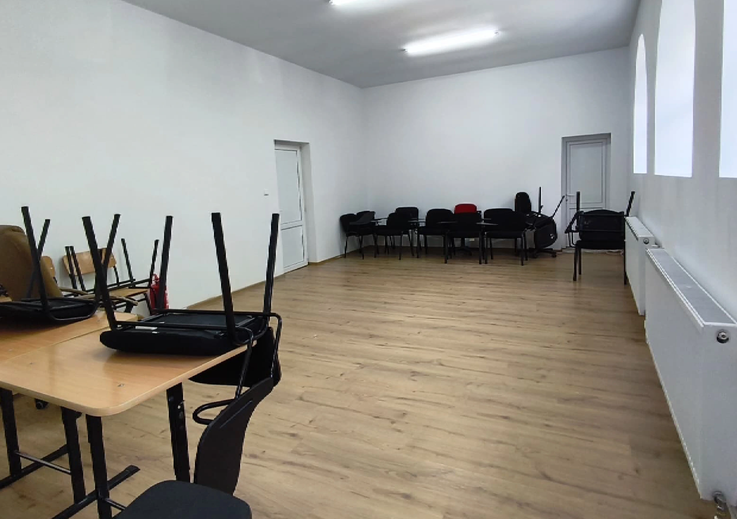 Sală renovată, cu pardoseală din lemn deschis, pereți albi și scaune stivuite lângă perete.